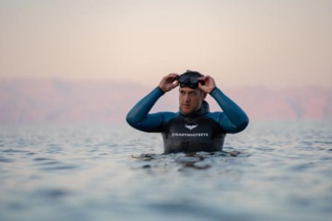 A white man in a wetsuit standing in the sea adjusting his goggles 