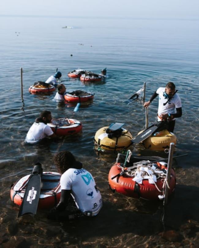 Several people in wetsuits under T-shirts at the water’s edge push floats out to sea 
