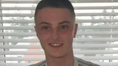 GoFundMe John Burns, standing in front of a window with open blinds, he is smiling and has shaved brown hair. 